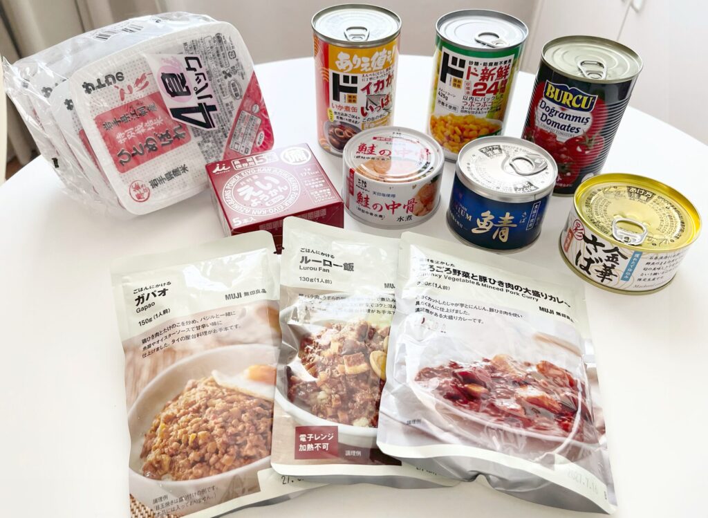 レトルトパック、ドンキホーテで購入したコーンやホールトマト、イカ、栄養缶、鮭の中骨、サバ缶、サバ味噌、金華サバ缶、無印良品のレトルトカレー、ルーロー飯、ガパオのレトルトパック商品を机の上に並べた